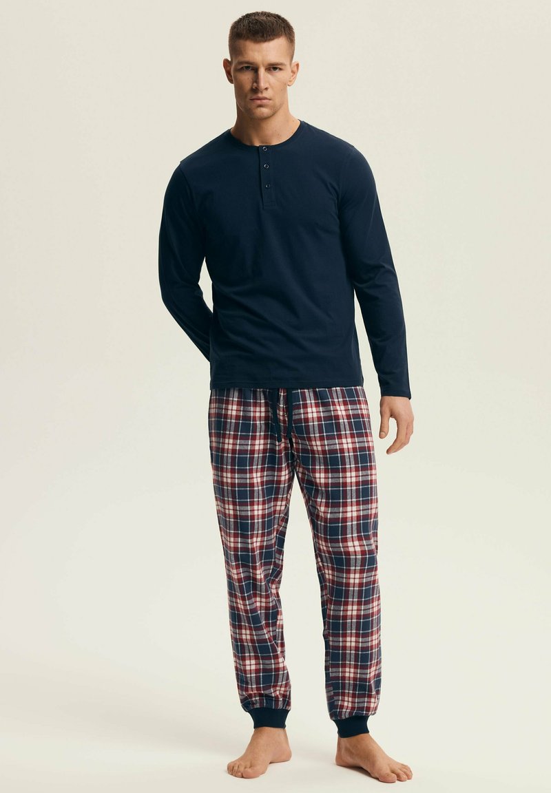 Mężczyzna w granatowej, długorękawowej koszulce henley i czerwono-biało-granatowych flanelowych spodniach do piżamy, stojący boso na beżowym tle.