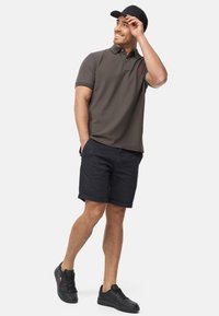 Camisa polo de oliva oscura con acentos rojos, pantalones cortos negros y zapatillas negras. El modelo está de pie de manera casual con una mano en el bolsillo, sonriendo.