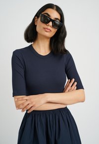 Marineblaue gerippte Crop-Top mit kurzen Ärmeln, kombiniert mit einem fließenden, dunklen Rock. Das Model trägt auffällige schwarze, rechteckige Sonnenbrillen.
