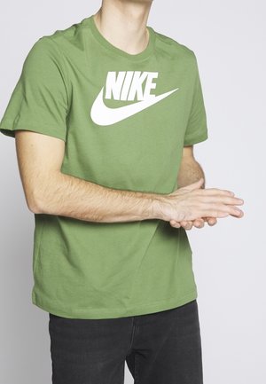 Persoon met een groen Nike T-shirt en zwarte broek, die de handen voor zich samenknijpt voor een effen lichte achtergrond.