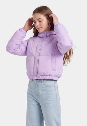 Junge Frau richtet ihr langes Haar, trägt eine hellviolette Steppjacke und hellblaue Jeans mit hoher Taille vor weißem Hintergrund.