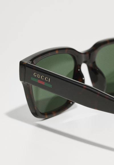 Nahaufnahme von dunkelbraunen Gucci-Sonnenbrillen mit grünen Gläsern und Gucci-Logo in Gold am Bügel.