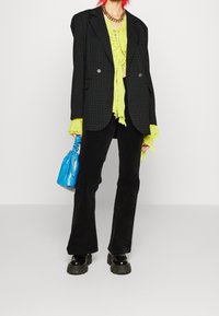 Černý kostkovaný oversized blazer, neonově zelený vrstvený top, černé zvonové kalhoty, masivní černé platformové boty a malá modrá kabelka.