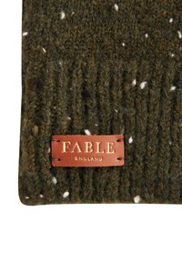 Zaļš adīts cepure ar teksturētu rakstu un baltām svītrām. Uz tā ir iešūta brūna ādas etikete ar iegravētu tekstu.