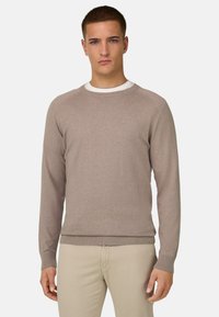 Jeune homme portant un pull beige à col rond par-dessus une chemise blanche et un pantalon beige clair, debout devant un fond clair uni.