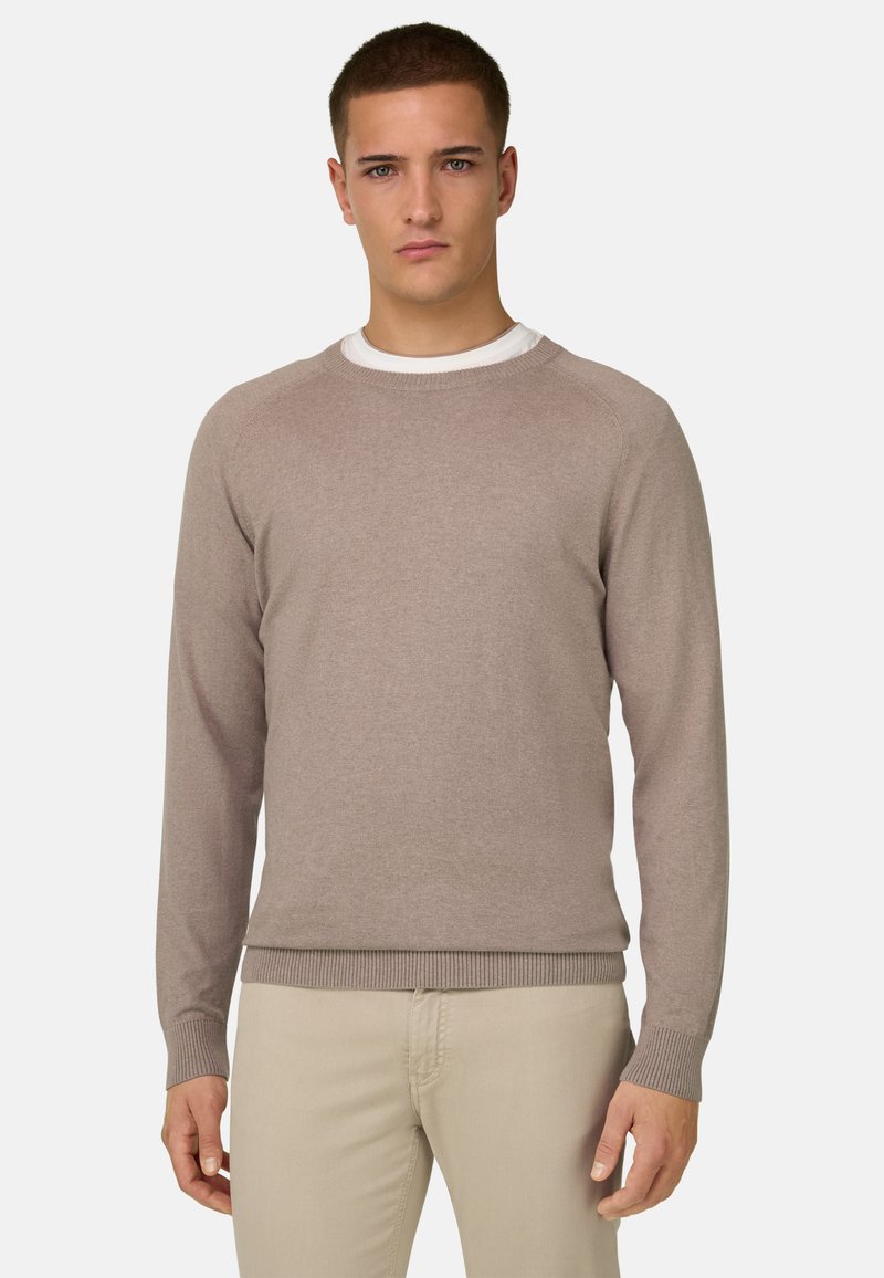 Jeune homme portant un pull beige à col rond par-dessus une chemise blanche et un pantalon beige clair, debout devant un fond clair uni.