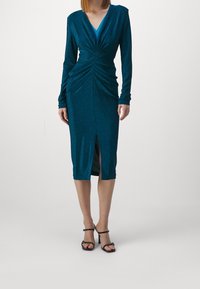 Tealfärgad glittrande knälång klänning med långa ärmar, V-ringning och rynkor framtill, med en hög slits vid låret och figurnära silhuett.