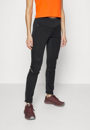 Person iført sorte, tætsiddende træningsbukser med lynlåslommer, orange ærmeløs top og bordeaux løbesko, stående på en ensfarvet hvid gulv.