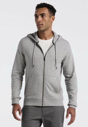 Mann trägt eine hellgraue Kapuzenjacke mit Reißverschluss, Kordelzug und Vordertaschen, kombiniert mit dunkelgrauen Hosen, blickt leicht nach links.