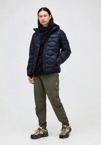 Black puffer jacket with quilted pattern, hood, and front zip. Worn over a black long-sleeve top, paired with olive green pants and hiking shoes.