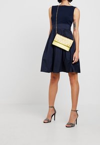 Námořnické bezramínkové šaty s těsným korzetem a rozšířenou sukní; doplněné žlutou prošívanou kabelkou a černými sandály na podpatku s třpytivými akcenty.