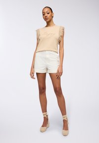 Top beige chiaro con maniche a frange, con la scritta "Enjoy" in corsivo. Pantaloni corti bianchi con risvolti. Zeppole beige in espadrille con lacci alla caviglia.