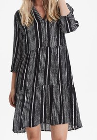 Musta-valkoinen raidallinen mekko, jossa on V-pääntie, kolmiviidesosaiset hihat ja kerroksellinen, levenevä helma. Kangas näyttää kevyeltä ja rakenteelliselta.