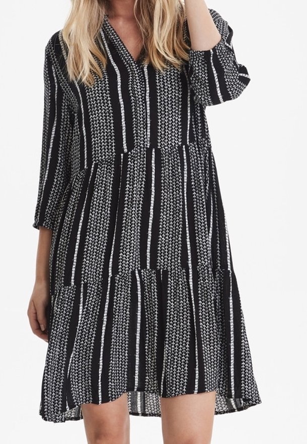 Musta-valkoinen raidallinen mekko, jossa on V-pääntie, kolmiviidesosaiset hihat ja kerroksellinen, levenevä helma. Kangas näyttää kevyeltä ja rakenteelliselta.