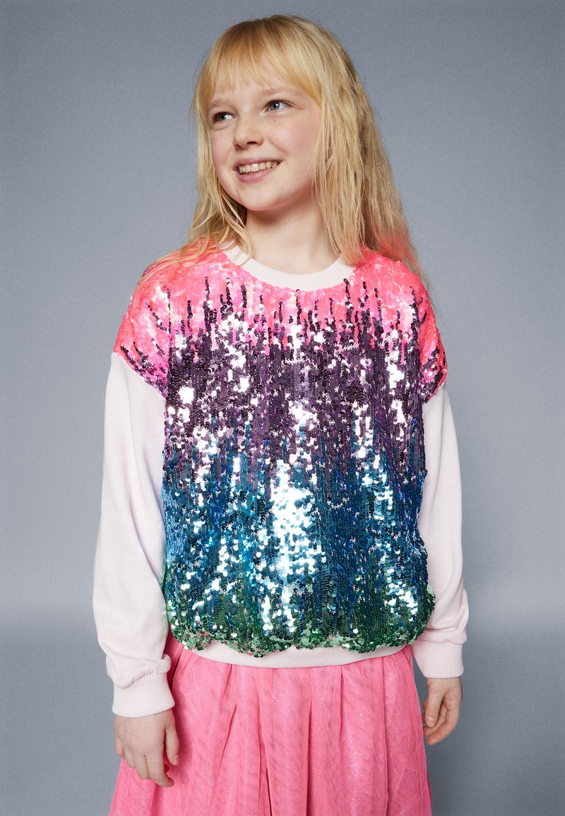 Svetra sa šljokicama koji prikazuje gradijent ružičastih, ljubičastih, plavih i zelenih šljokica. Bele rukave i rebrasti izrez. Uparen s ružičastom suknjom.