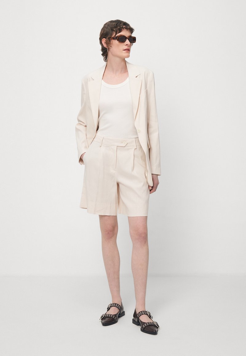 Hellbeige taillierter Blazer über einem weißen Tanktop, kombiniert mit passenden Shorts. Kleine Taschen, glattes Material und schwarzen Sandalen mit Blockabsatz und Akzenten.