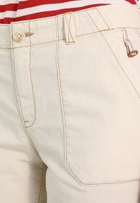 Pantalones de algodón beige con una textura suave, que presentan un cierre de botón y costuras en contraste. El diseño incluye bolsillos frontales.