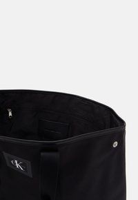 Black tote bag made of nylon, featuring a smooth texture. Interior includes a zippered pocket and a logo patch. Top opening with a simple design.
