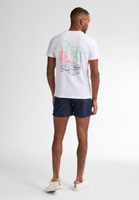 Tricou scurt cu mânecă albă, având un grafic colorat cu palmieri și un camion vintage pe spate, asortat cu pantaloni şort bleumarin și adidași deschiși la culoare.