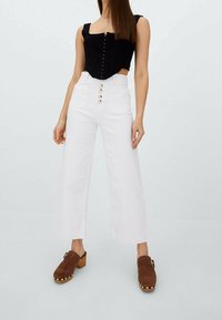 Top corto negro texturizado con escote cuadrado y cierre de botones, combinado con pantalones anchos blancos que presentan un detalle de botón en la parte delantera. Zuecos marrones.