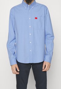 Camisa de botones de tejido azul claro con una textura suave. Presenta un parche de logo rojo en el bolsillo del pecho y un cuello. Mangas largas.