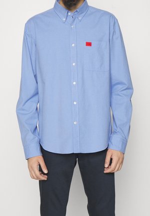 Button-up shirt in light blue fabric with a smooth texture. Features a red logo patch on the chest pocket and a collar. Long sleeves.