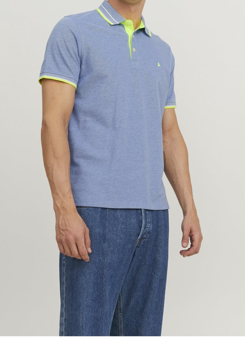Homme portant un polo bleu clair à manches courtes avec un col et un bord de manches jaune néon, associé à un jean bleu, debout les bras détendus.
