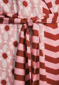 Raštuotas audinys, kuriame pakaitomis yra rožinės ir raudonos geometrinės formos su surišamu juosmeniu. Spaudinys apima juosteles ir formas.