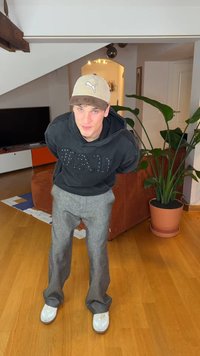 Must kapuutsiga pusa tekstuurse kirjaga, helehall ruuduline püksid ja valged tossud. Taustal on taim ja pruun diivan.