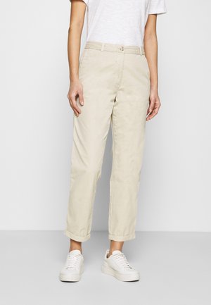 Persona con pantalones beige con puños, zapatillas blancas y una camisa blanca de manga corta, de pie frente a un fondo claro y sencillo.