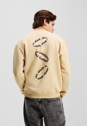 Beige Sweatshirt aus strukturiertem Stoff mit runder schwarzer Schrift auf der Rückseite. Das Design umfasst den Markennamen und grafische Elemente.