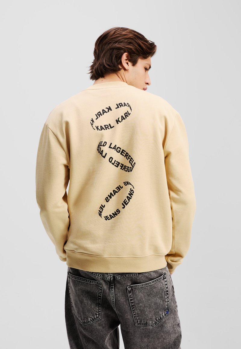 Sweatshirt beige en tissu texturé avec des lettres noires rondes au dos. Le design inclut le nom de la marque et des éléments graphiques.