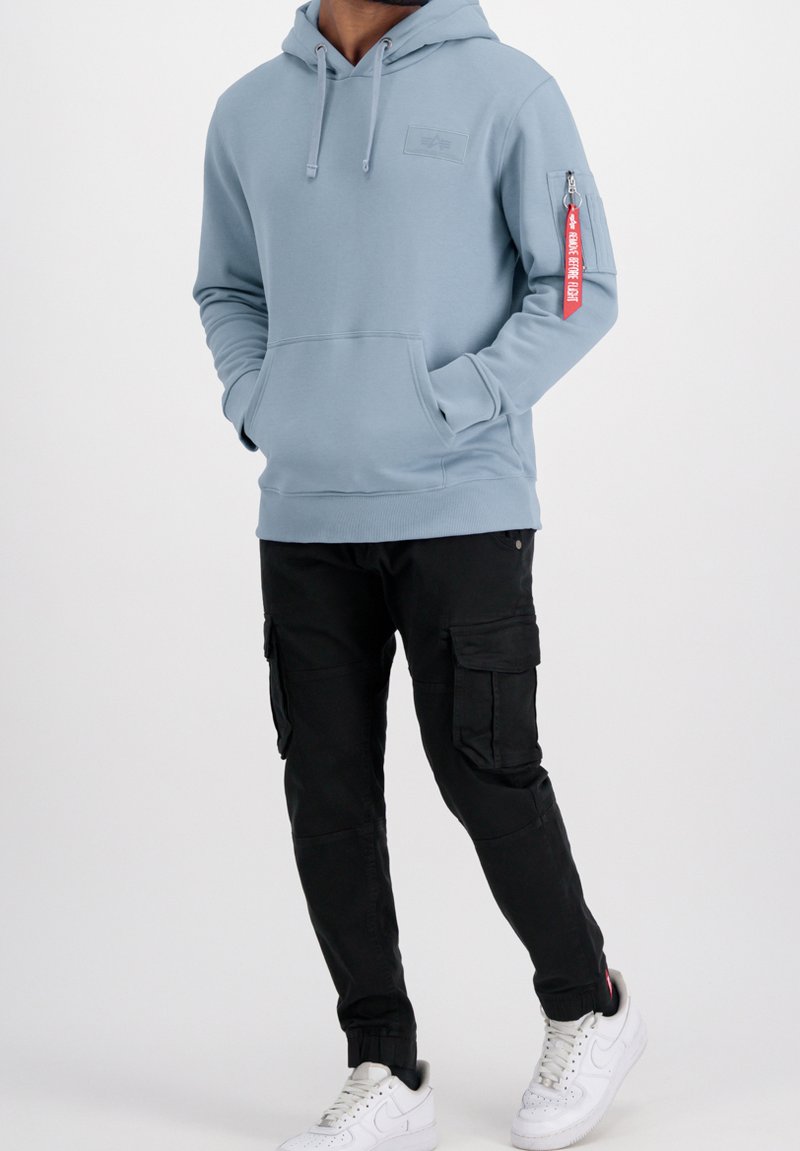 Svijetloplava dukserica s prednjim džepom, kapuljačom na vezicu i džepom na rukavu sa zatvaračem s crvenim detaljem. U kombinaciji s crnim cargo hlačama i bijelim tenisicama.