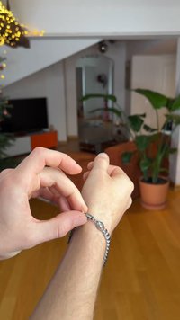 Μεταλλικό βραχιόλι με διασυνδεδεμένους κρίκους και κούμπωμα, που δένεται σε καρπό. Το φόντο περιλαμβάνει φυτά εσωτερικού χώρου και έπιπλα.