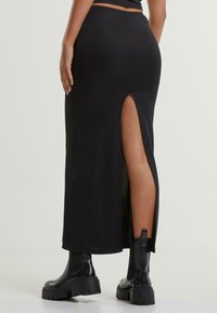 Schwarzer Maxi-Rock mit seitlichem Schlitz, aus einem weichen Stoff gefertigt. Kombiniert mit klobigen schwarzen Stiefeletten, die eine schlanke, taillierte Silhouette betonen.