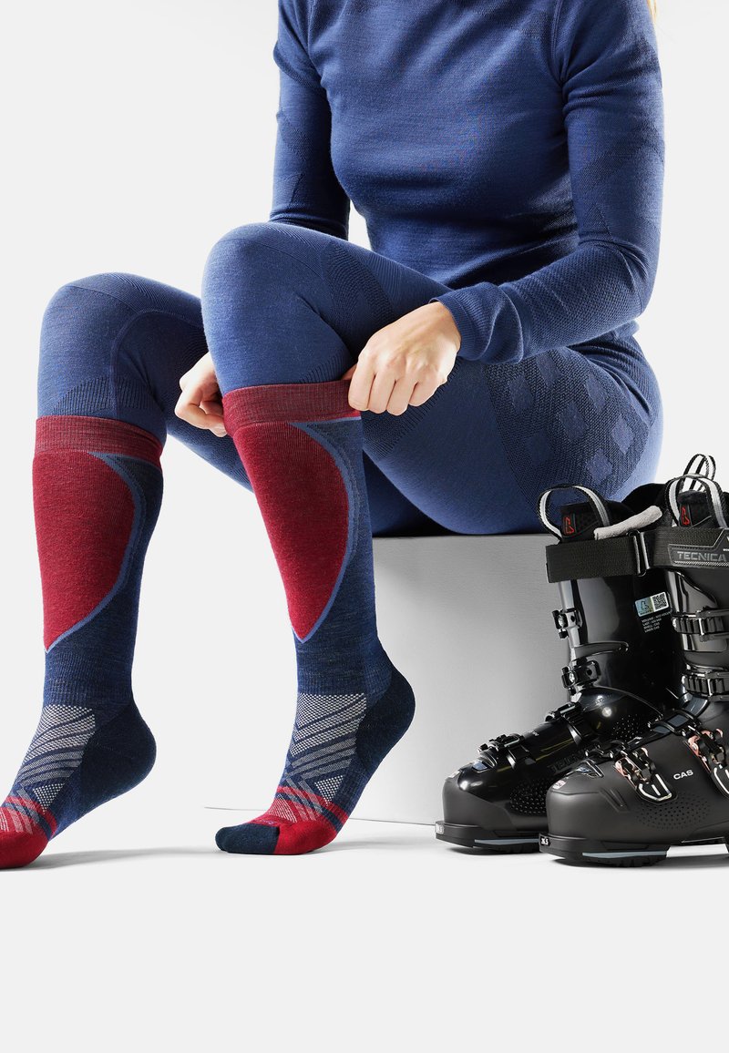 Skisokken in blauw en rood met patroonaccenten; liggen naast zwarte skibotten. Het materiaal lijkt synthetisch met een ribbelstructuur.