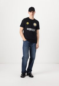 T-shirt en coton noir avec des logos jaunes et blancs. Associé à un jean en denim bleu et des chaussures noires. Le modèle porte une casquette noire.