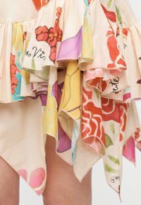 Αέρινη φούστα με στρωματοποιημένα, μαντηλοειδή τελειώματα που φέρουν πολύχρωμα λουλουδάτα σχέδια σε κόκκινο, ροζ, κίτρινο και μωβ, πάνω από γυμνά πόδια.