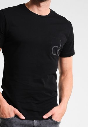 Mężczyzna ubrany w czarny t-shirt z krótkim rękawem z małą kieszenią po lewej stronie na klatce piersiowej z szarym logo "ck", ręce w kieszeniach.
