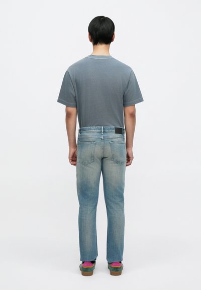 Γκρι κοντομάνικο μπλουζάκι, ανοιχτό μπλε τζιν με ξεθώριασμα, ίσια γραμμή παντελονιού και καφέ παπούτσια με πράσινες και ροζ λεπτομέρειες. Απλό φόντο.