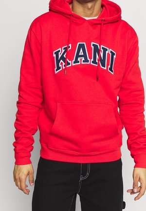 Person trägt einen leuchtend roten Hoodie mit dem Schriftzug "KANI" in Schwarz und Weiß auf der Brust, kombiniert mit schwarzen Hosen mit weißen Nähten.