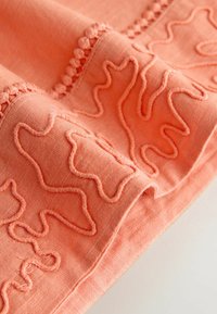 Tissu corail avec des motifs brodés ondulés en relief et un détail de bordure en pompons texturés le long du bord.