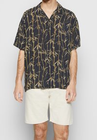 Camicia a maniche corte in tessuto blu navy con motivo in bambù verde e beige. Presenta un colletto e chiusura con bottoni, abbinata a pantaloni corti bianchi.
