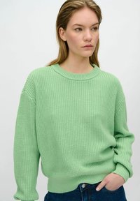 Hellgrüner Strickpullover mit Rundhalsausschnitt und gerippten Bündchen, lockere Passform und strukturierter Stoff. Kombiniert mit blauen Jeans.