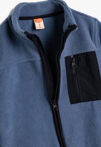 Fleecejacke in Hellblau mit einer schwarzen Reißverschlusstasche auf der Brust, hohem Kragen und einem schwarzen Reißverschluss vorne. Strukturierter, weicher Stoff.
