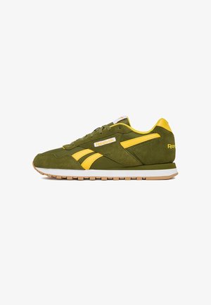 Zapatilla deportiva de ante verde con detalles en amarillo y suela intermedia blanca. Presenta franjas laterales contrastantes y logo de la marca en la lengüeta y el talón.