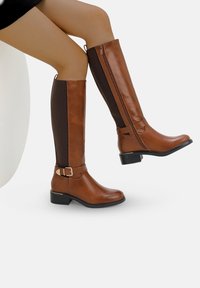 Botas marrones hasta la rodilla, hechas de cuero sintético liso, con paneles laterales elásticos, un cierre lateral y un acento de hebilla en el tobillo.