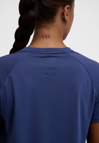 Niebieska koszulka sportowa z teksturowanej tkaniny. Posiada subtelny wzór z trzema strzałkami skierowanymi w dół oraz tatuażem z napisem "444" na szyi.