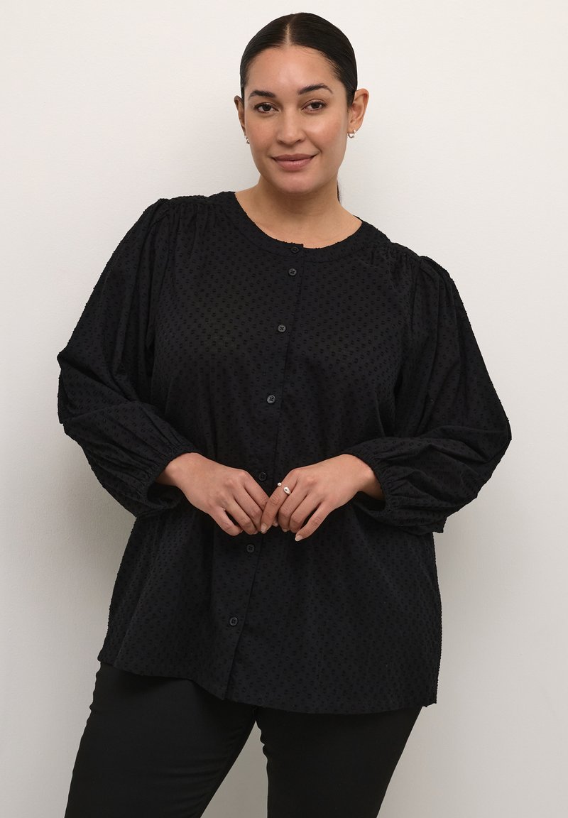 Camicia nera con maniche a sbuffo, tessuto testurizzato, scollo rotondo e sette bottoni sul davanti, abbinata a pantaloni neri.