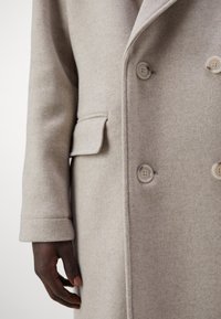 Manteau en laine beige clair à double boutonnage, avec deux boutons et une poche latérale. Texture douce et coupe ajustée.